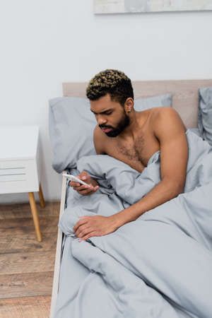 shirtless african american man with dyed hair messaging on smartphone in bedの写真素材