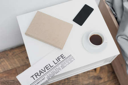 top view of cup with coffee and smartphone near newspaper with travel life lettering and book on white bedside tableの写真素材