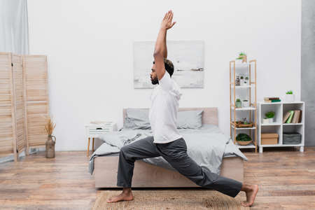 side view of bearded african american man practicing yoga in warrior pose and meditating in bedroom