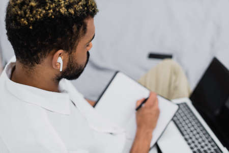 young african american man in wireless earphone using laptop while writing in notebook