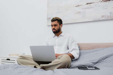 young african american freelancer in wireless earphone using laptop while sitting on bed at homeの写真素材