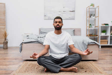 bearded african american man in wireless earphone meditating with closed eyes while sitting on rug...