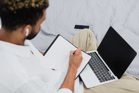 high angle view of blurred african american man in wireless earphone using laptop while writing in...