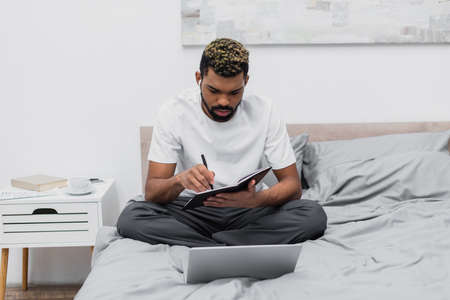 bearded african american man in wireless earphones looking at notebook near laptop on bed