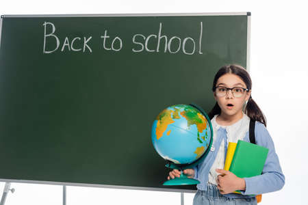 Shocked schoolkid holding books and globe near chalkboard with back to school lettering isolated on whiteの写真素材