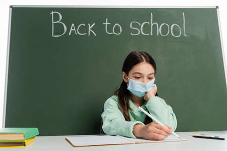 Preteen pupil in medical mask writing on notebook near chalkboard with back to school lettering...