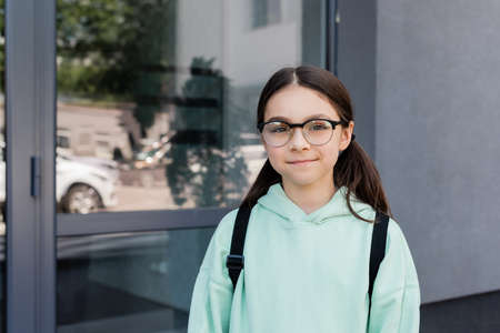 Smiling schoolgirl in eyeglasses looking at camera near building outdoorsの写真素材