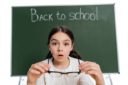Shocked schoolchild holding eyeglasses near blurred chalkboard with back to school lettering isolated on whiteの写真素材