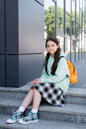 Schoolkid with backpack looking at camera on stairs near building on urban streetの写真素材