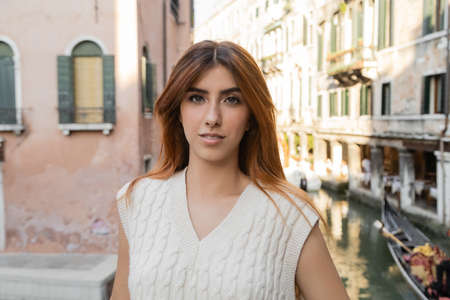 portrait of pretty woman in sleeveless jumper looking at camera near blurred venetian buildingsの写真素材