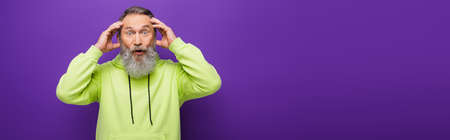 shocked and bearded senior man with grey hair looking at camera on purple, bannerの写真素材