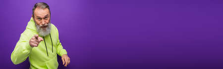 senior man with beard and grey hair pointing with finger at camera on purple, banner