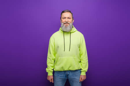 cheerful bearded man with grey hair and green hoodie smiling on purpleの写真素材