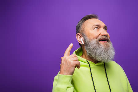 happy senior man in green hoodie listening music and pointing with finger at wireless earphone on purpleの写真素材