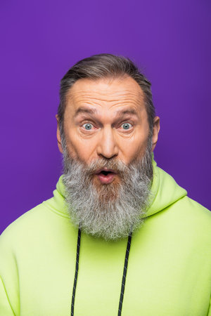 astonished senior man opening mouth while looking at camera on purpleの写真素材