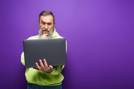 pensive senior man with beard and grey hair using laptop on purple background