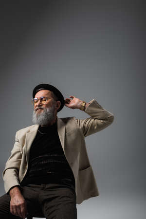 bearded senior man in eyeglasses and jacket adjusting derby hat on grey