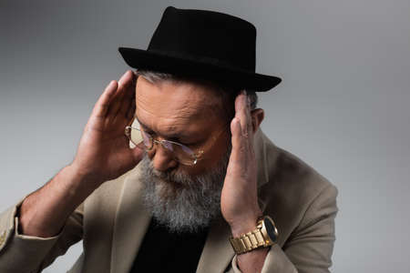 bearded senior man in derby hat and eyeglasses looking down on grey