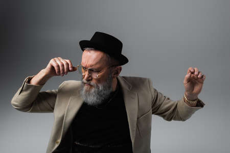 bearded senior man in derby hat and eyeglasses gesturing on greyの写真素材