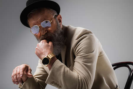 portrait of bearded senior man in stylish beige jacket and derby hat looking at camera on greyの写真素材