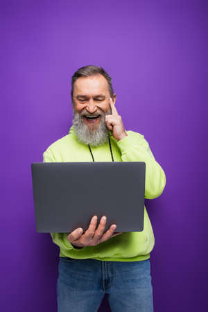 positive senior man with beard and grey hair using laptop on purpleの写真素材
