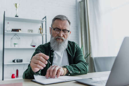 bearded senior man holding pen and looking at notebook near laptop on desk