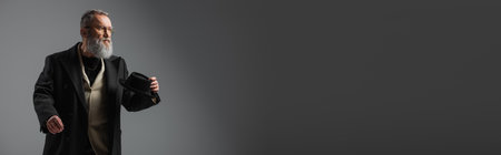 elegant senior man in stylish coat and eyeglasses holding derby hat on grey, bannerの写真素材