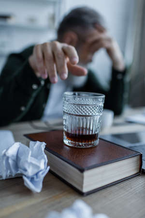 blurred senior man reaching glass of whiskey on book near crumpled paperの写真素材