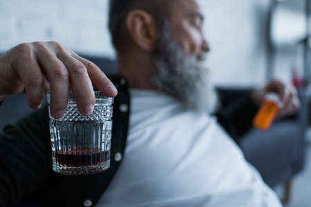 blurred and unhappy senior man holding glass with whiskey and bottle with medication at homeの写真素材