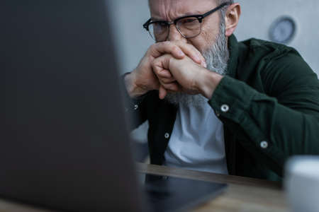 stressed senior man in eyeglasses frowning and looking at blurred laptop at homeの写真素材