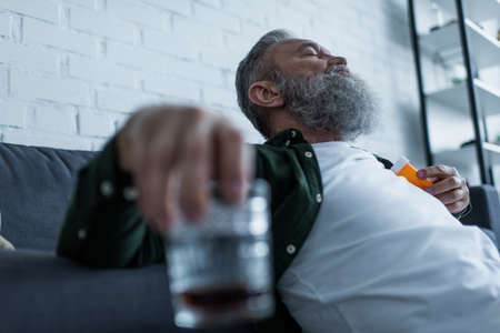 bearded senior man with grey hair holding bottle with medication and glass of whiskeyの写真素材