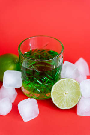 frozen ice cubes in glass with alcohol green beverage and limes on red