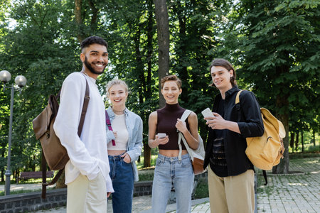 Smiling interracial students with backpacks and smartphones looking at camera in park