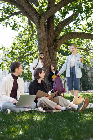 Cheerful student with takeaway drink standing near multiethnic friends on grass in summer parkの写真素材