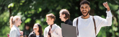 Excited african american student holding laptop near blurred friends in summer park, bannerの写真素材