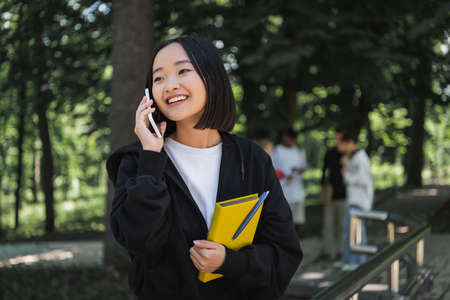 Positive asian student holding book and talking on smartphone in park