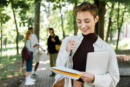 Cheerful student with laptop looking at notebook in summer parkの写真素材