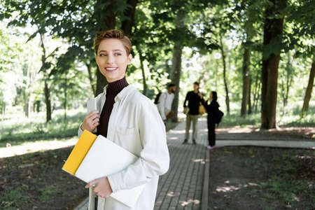 Cheerful student holding gadgets and notebook in summer park