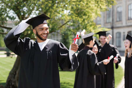 Cheerful african american bachelor looking at diploma near blurred friends in parkの写真素材
