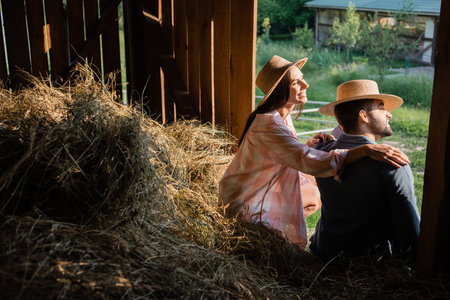 happy woman in straw hat hugging shoulders of husband while sitting on haystack in barnの写真素材