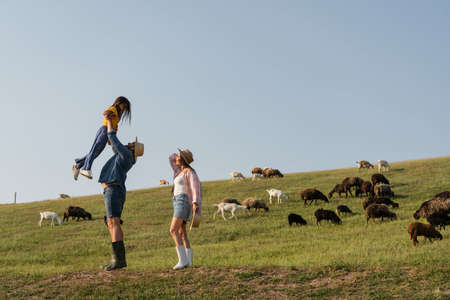 side view of farmer raising up daughter near wife and herd grazing in green pastureの写真素材