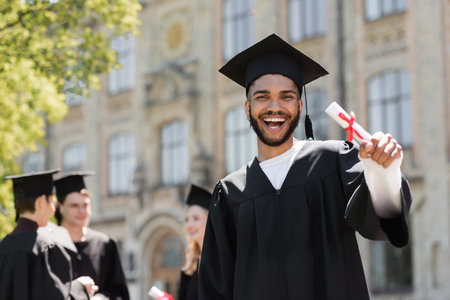 Happy african american bachelor in gown holding diploma outdoorsの写真素材