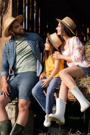 cheerful couple in straw hats looking at each other near daughter on hay in barnの写真素材