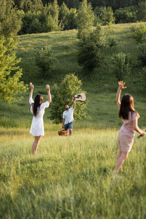 mom and daughter waving hands to man with guitar on picturesque slopeの写真素材