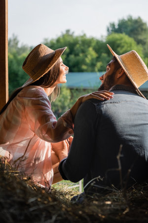 joyful woman in straw hat touching shoulder of husband near blurred hey in barnの写真素材