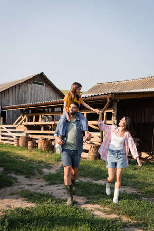 cheerful man piggybacking daughter while walking near wife on rural farmの写真素材