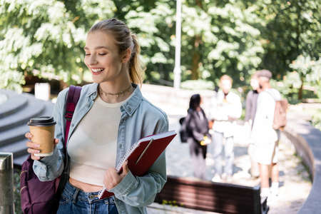 Smiling student holding coffee to go and notebook in parkの写真素材