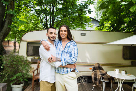 happy gay couple smiling while hugging near travel van in forest