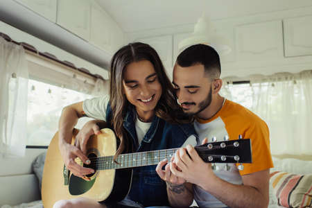smiling gay man with long hair playing acoustic guitar near happy boyfriend in modern vanの写真素材