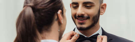 tattooed gay man adjusting bow tie on suit of happy bearded groom, banner